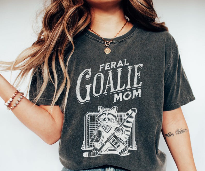 May include: A dark gray t-shirt with a white graphic of a raccoon wearing a hockey goalie uniform and the text "Feral Goalie Mom".