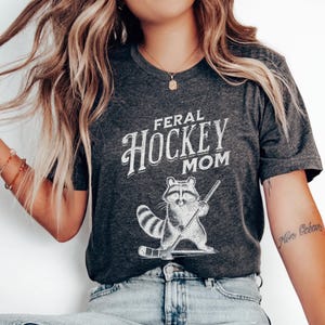 May include: A dark gray t-shirt with the text "Feral Hockey Mom" in white lettering. A cartoon raccoon holding a hockey stick is printed below the text.