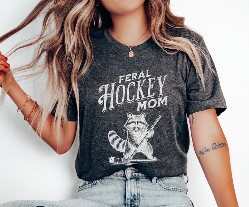 May include: A dark gray t-shirt with the text "Feral Hockey Mom" in white lettering. A cartoon raccoon holding a hockey stick is printed below the text.