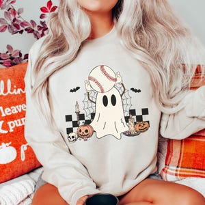 May include: A white sweatshirt with a graphic of a ghost holding a baseball. The ghost is surrounded by bats, a spiderweb, candles, and pumpkins. The text "Boo" is printed on the sweatshirt.