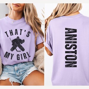 May include: A lavender t-shirt with a hockey player graphic and the text "That's My Girl" on the front. The back of the shirt has the text "ANISTON" in a distressed font.