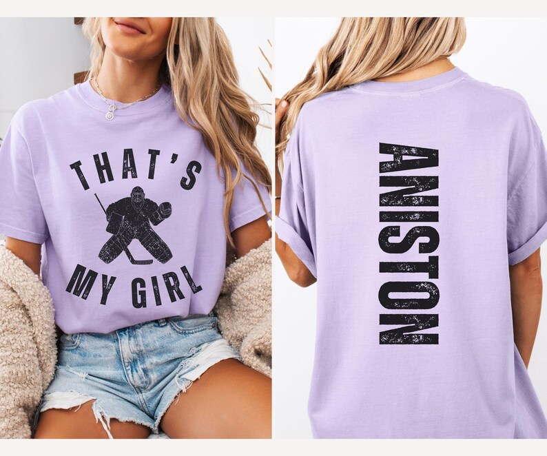May include: A lavender t-shirt with a hockey player graphic and the text "That's My Girl" on the front. The back of the shirt has the text "ANISTON" in a distressed font.