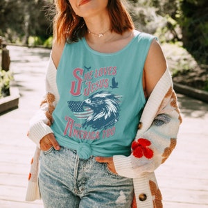 May include: A woman wearing a light blue tank top with the text "She Loves Jesus and America Too" in red, white, and blue. The tank top features a graphic of an eagle with an American flag in the background.