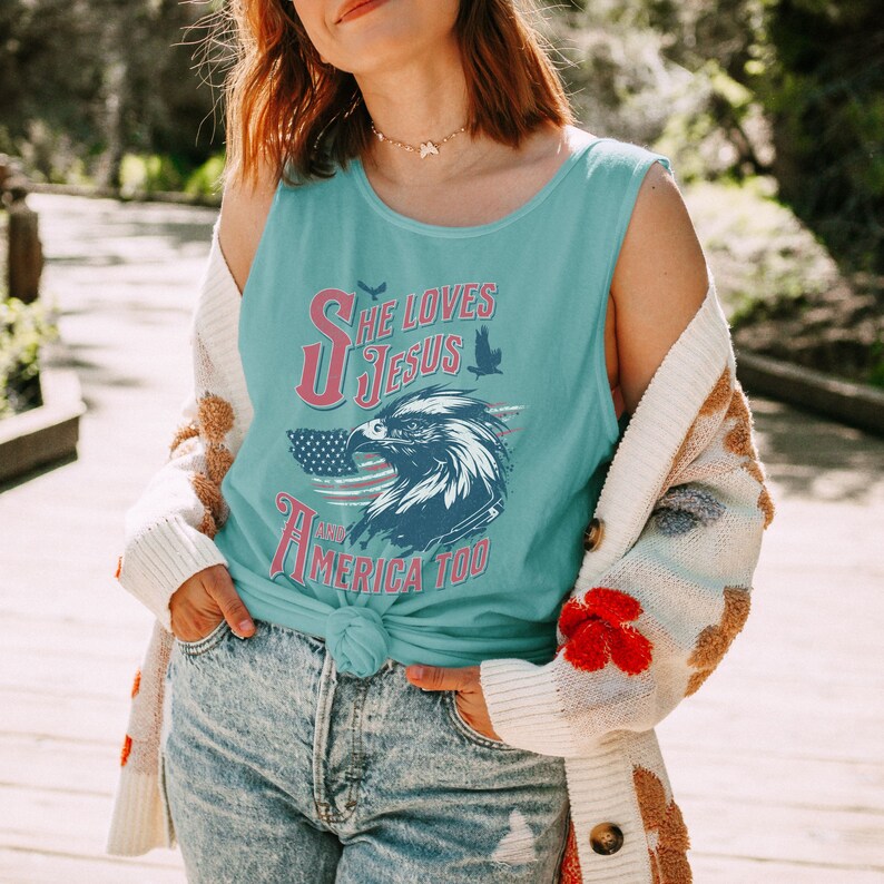 May include: A woman wearing a light blue tank top with the text "She Loves Jesus and America Too" in red, white, and blue. The tank top features a graphic of an eagle with an American flag in the background.
