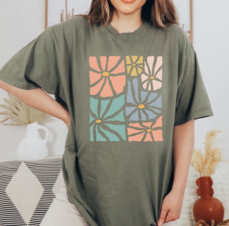 Op de afbeelding: Een groen T-shirt met een grafische print van kleurrijke abstracte bloemen. De bloemen zijn in een vierkant patroon gerangschikt en hebben een retro stijl.