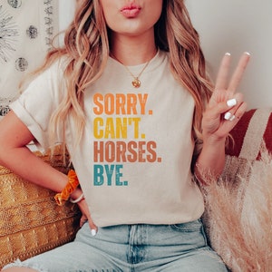 May include: A woman wearing a light beige t-shirt with the text "SORRY. CAN'T. HORSES. BYE." printed in a vintage style with orange, yellow, blue, and green colors.
