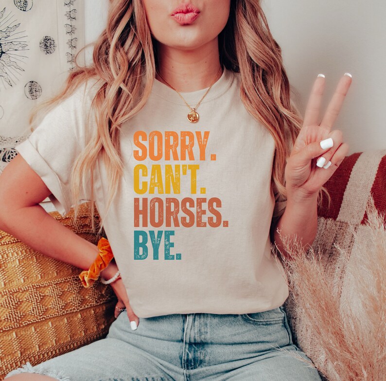 May include: A woman wearing a light beige t-shirt with the text "SORRY. CAN'T. HORSES. BYE." printed in a vintage style with orange, yellow, blue, and green colors.