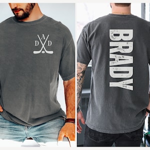 Puede incluir: Una camiseta gris con un dise&ntilde;o de palo de hockey blanco en la parte delantera y la palabra "BRADY" en letras blancas desgastadas en la parte trasera.