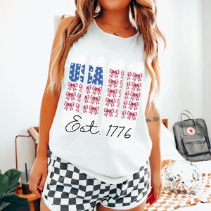 May include: White vest top with a USA flag design featuring blue stars and red bows. The text "Est. 1776" is printed below the flag design. The model is wearing black and white checkered shorts.