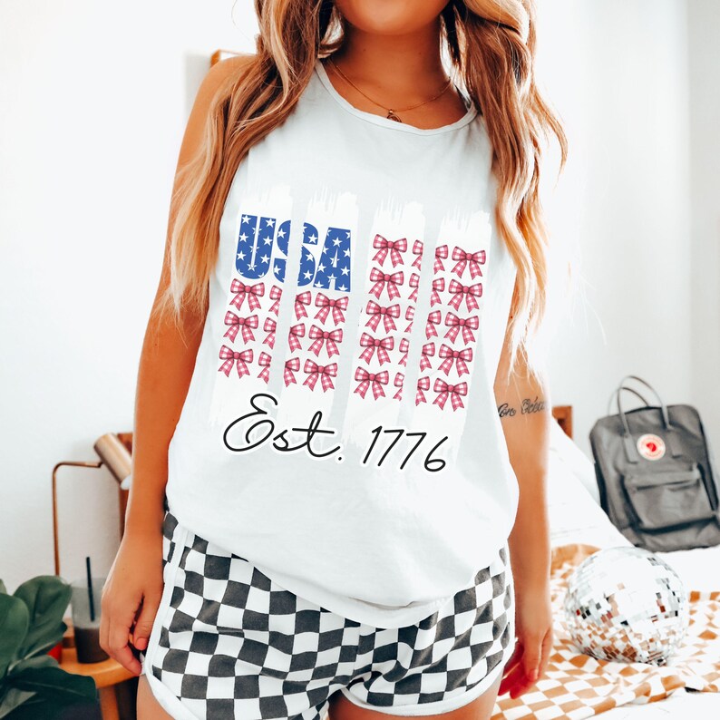 May include: White vest top with a USA flag design featuring blue stars and red bows. The text "Est. 1776" is printed below the flag design. The model is wearing black and white checkered shorts.