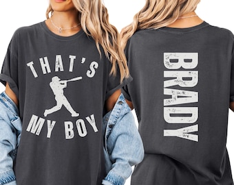 May include: A charcoal gray t-shirt featuring a white distressed graphic on the front with a baseball player silhouette and the text THAT'S MY BOY. The back of the shirt displays the name BRADY in large, vertical, white distressed lettering.