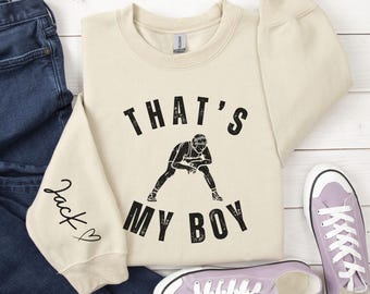 Puede incluir: Una sudadera beige con el texto "That's My Boy" en negro. Una silueta de un luchador está en el centro del texto. La manga izquierda tiene el nombre "Jack" escrito en cursiva con un corazón.