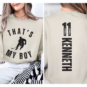 Może przedstawiać: Beżowa bluza z czarnym nadrukiem hokeisty i tekstem "That's My Boy" z przodu. Z tyłu bluzy znajduje się numer "11" i imię "Kenneth" pionowo na plecach.
