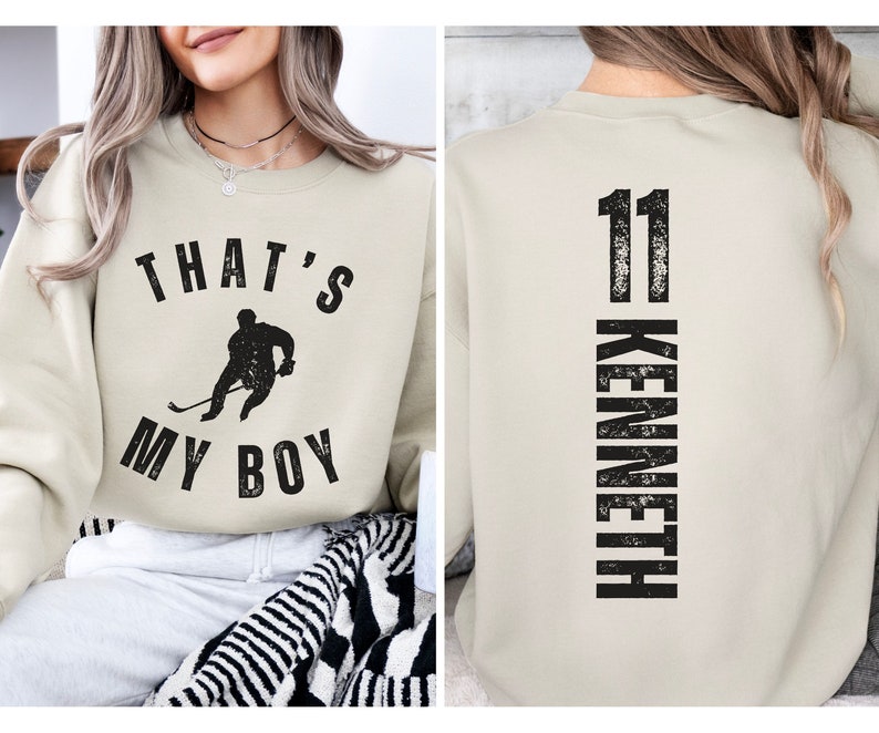 Może przedstawiać: Beżowa bluza z czarnym nadrukiem hokeisty i tekstem "That's My Boy" z przodu. Z tyłu bluzy znajduje się numer "11" i imię "Kenneth" pionowo na plecach.