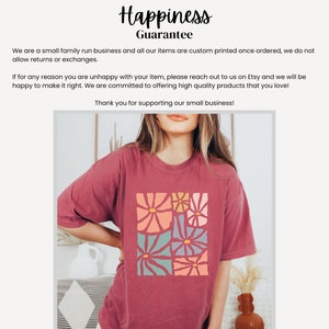 Op de afbeelding: Een maron t-shirt met een kleurrijk bloemenontwerp dat op de voorkant is gedrukt. Het ontwerp heeft een vierkant patroon van bloemen in de kleuren roze, geel, blauw en groen.