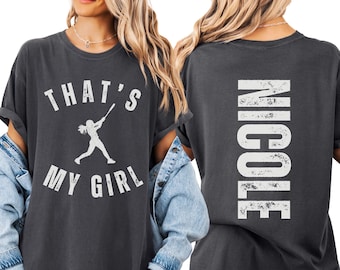 May include: A charcoal gray t-shirt featuring a white distressed graphic on the front with a silhouette of a softball player swinging a bat and the text THAT'S MY GIRL. The back of the shirt displays the name NICOLE in large white vertical letters.