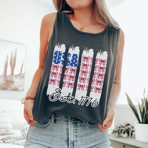 May include: A dark grey vest top with a patriotic design. The design features the word "USA" in blue with white stars, and red and white bows. The text "Est. 1776" is below the design.
