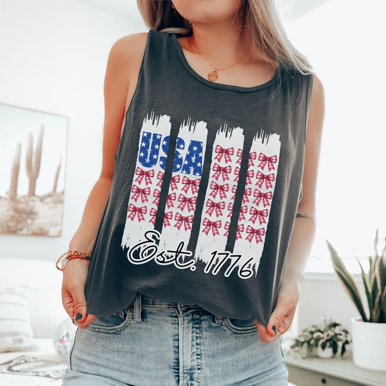 May include: A dark grey vest top with a patriotic design. The design features the word "USA" in blue with white stars, and red and white bows. The text "Est. 1776" is below the design.