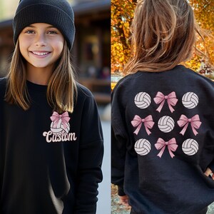 May include: Black sweatshirt with a volleyball and pink bow design on the front and a pattern of volleyballs with pink bows on the back. The text "Custom" is on the front of the sweatshirt.