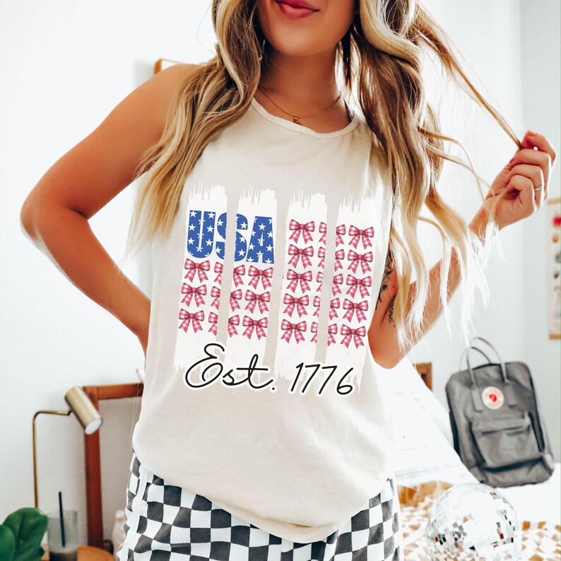 May include: Cream-coloured tank top with a patriotic design. The design features the word "USA" in blue stars and stripes, along with rows of red and pink bows. The text "Est. 1776" is written in a cursive font.