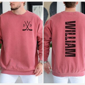 Puede incluir: Una sudadera roja con un dise&ntilde;o de palo de hockey negro en la parte delantera y la palabra "WILLIAM" impresa verticalmente en la parte posterior.