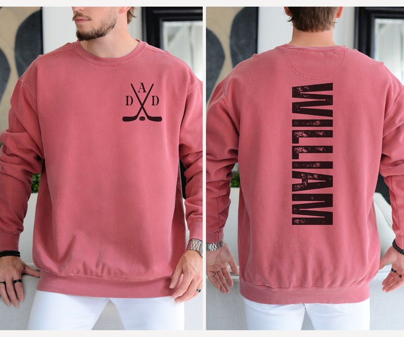 Puede incluir: Una sudadera roja con un dise&ntilde;o de palo de hockey negro en la parte delantera y la palabra "WILLIAM" impresa verticalmente en la parte posterior.