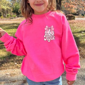 May include: A bright pink sweatshirt with a volleyball graphic and a pink bow. The text "Kylie" is printed in a retro font below the volleyball.