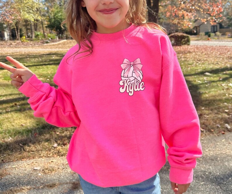 May include: A bright pink sweatshirt with a volleyball graphic and a pink bow. The text "Kylie" is printed in a retro font below the volleyball.