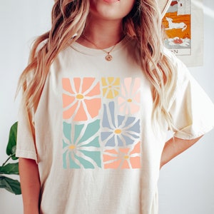 Op de afbeelding: Een cr&egrave;mekleurig T-shirt met een grafische print van pastelkleurige bloemen. De bloemen zijn in een vierkant patroon gerangschikt en hebben een retro-stijl.