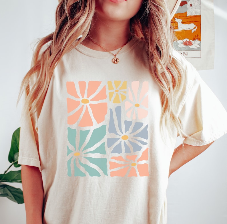 Op de afbeelding: Een cr&egrave;mekleurig T-shirt met een grafische print van pastelkleurige bloemen. De bloemen zijn in een vierkant patroon gerangschikt en hebben een retro-stijl.