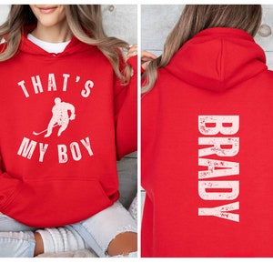 Może przedstawiać: Czerwona bluza z kapturem z białym nadrukiem, kt&oacute;ry głosi "That's My Boy" z sylwetką hokeisty. Tył bluzy ma biały nadruk, kt&oacute;ry głosi "BRADY" w stylu vintage.
