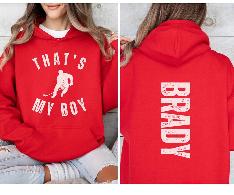Może przedstawiać: Czerwona bluza z kapturem z białym nadrukiem, kt&oacute;ry głosi "That's My Boy" z sylwetką hokeisty. Tył bluzy ma biały nadruk, kt&oacute;ry głosi "BRADY" w stylu vintage.