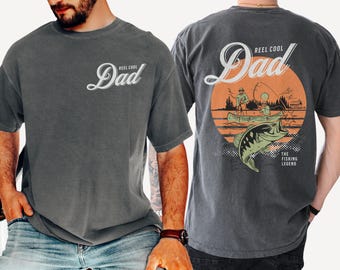 May include: A charcoal gray t-shirt featuring a small white graphic on the front that reads Reel Cool Dad. The back displays a large orange circular graphic with a fishing scene, a jumping bass, and the text Reel Cool Dad The Fishing Legend.