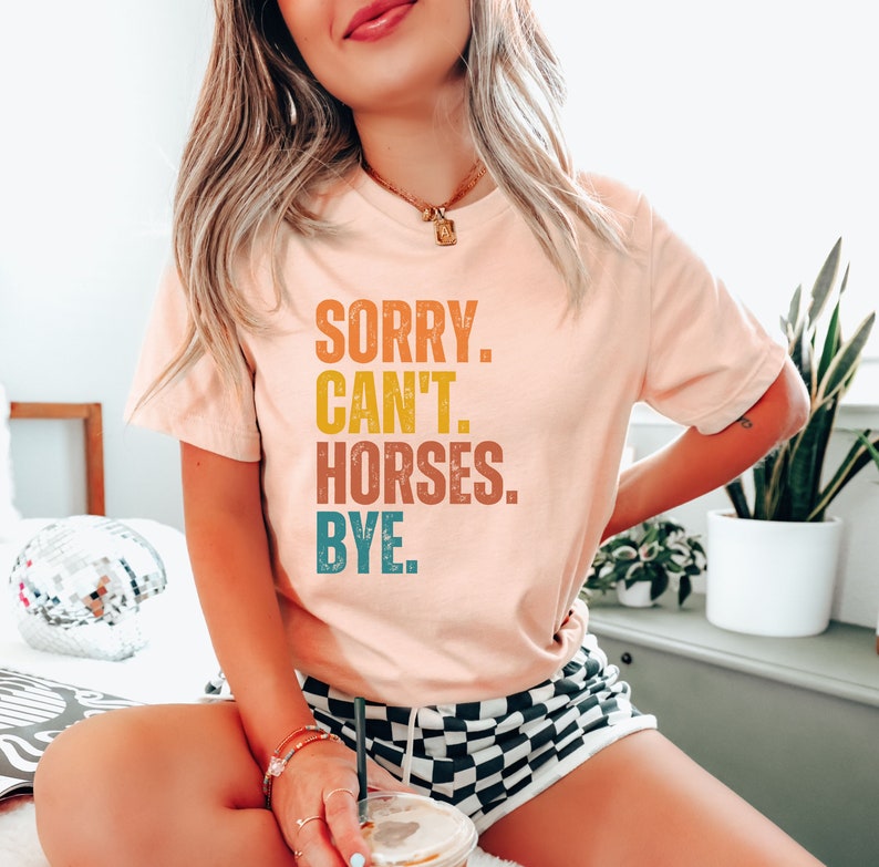 May include: A pink t-shirt with the text "SORRY. CAN'T. HORSES. BYE." in a vintage style font with a yellow, orange, and blue color scheme.