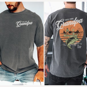 May include: A dark gray t-shirt with the text "Reel Cool Grandpa" and a graphic of a fish jumping out of the water. The graphic also includes a sunset, trees, and two people fishing in a boat. The text "The Fishing Legend" is below the graphic.