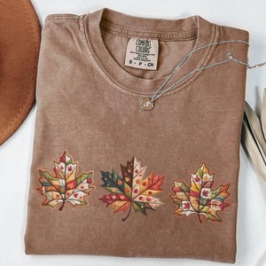 May include: A brown Comfort Colors t-shirt featuring three colorful maple leaf designs. The leaves are filled with patchwork patterns in shades of red, orange, yellow, and green. A silver necklace with a small pendant rests on the shirt.