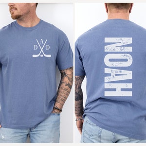 Puede incluir: Una camiseta azul con un dise&ntilde;o de palo de hockey blanco en la parte delantera y la palabra "NOAH" en blanco en la parte posterior.