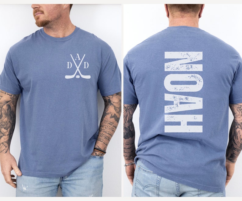 Puede incluir: Una camiseta azul con un dise&ntilde;o de palo de hockey blanco en la parte delantera y la palabra "NOAH" en blanco en la parte posterior.