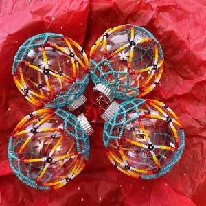 May include: Four clear glass spherical ornaments, each wrapped in a geometric web of small seed beads. The beadwork features a vibrant color palette of turquoise, yellow, orange, red, white, and black, arranged in a repeating triangular pattern.