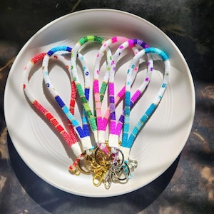 May include: A collection of beaded lanyards in various colors, including red, blue, green, pink, and purple. Each lanyard features a white beaded section and a metal clasp. The lanyards are arranged on a white plate.