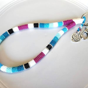 Pode incluir: Uma pulseira de chaveiro de contas em um gradiente de cores, incluindo roxo, branco, azul e preto. A pulseira é presa a um chaveiro de prata e a um mosquetão, com um pingente em forma de coração.