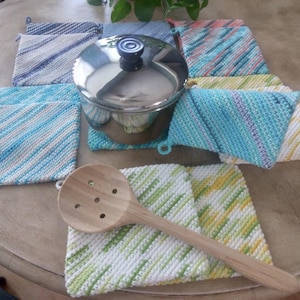 Op de afbeelding: Een verzameling kleurrijke, handgebreide pannenlappen in verschillende tinten blauw, groen, geel en roze. Een roestvrijstalen pan met een zwarte handgreep staat op een van de pannenlappen. Een houten lepel ligt op een pannenlap.