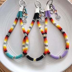 May include: Three beaded lanyards in turquoise, black, and purple, each with a silver keyring and a heart-shaped charm. The lanyards feature a colorful pattern of red, yellow, and white beads. The lanyards are displayed on a white plate.