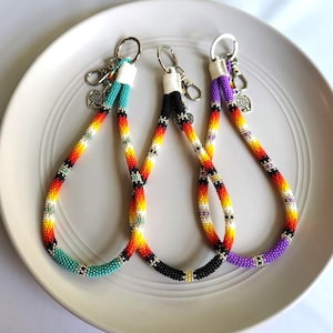 May include: Three beaded keychains in turquoise, black, and purple, each with a silver-tone clasp and charm. The keychains feature a colorful pattern of red, yellow, and white beads. They are displayed on a white plate.