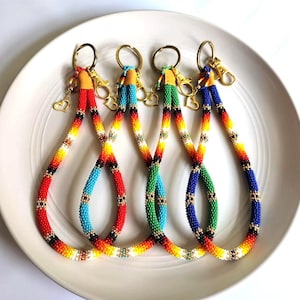 Puede incluir: Cuatro cordones con cuentas en rojo, turquesa, verde y azul, cada uno con un cierre dorado y un pequeño dije de corazón. Los cordones están dispuestos sobre un plato blanco, mostrando una vibrante exhibición de colores e intrincado trabajo de cuentas.