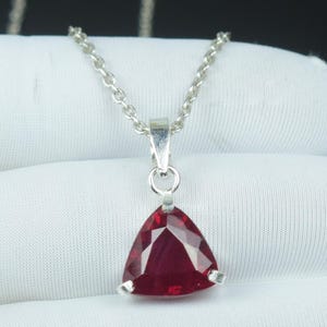 Puede incluir: Un collar de plata con un colgante triangular facetado de piedra preciosa rojo intenso. El colgante cuelga de una delicada cadena. La piedra preciosa está sujeta por tres garras de plata.