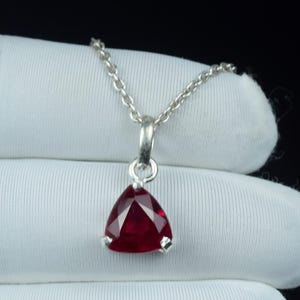 Op de afbeelding: Een zilveren ketting met een driehoekige, dieprode edelsteen hanger. De gefacetteerde hanger hangt aan een fijne zilveren ketting. De edelsteen is gezet in een zilveren zetting.