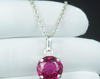 May include: A silver necklace featuring a round, bright pink gemstone pendant. The pendant is held in place by a four-prong setting and is suspended from a fine chain. The gemstone is a rich magenta hue.