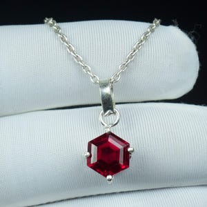 May include: A silver necklace featuring a hexagonal, deep red gemstone pendant. The pendant hangs from a delicate chain. The gemstone is faceted, reflecting light. The necklace is shown against a white backdrop.