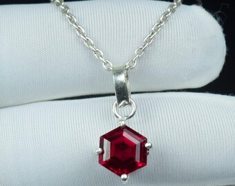 May include: A silver necklace with a hexagonal, deep red gemstone pendant. The pendant is suspended from a delicate chain. The gemstone is faceted, catching the light. The necklace is displayed against a white background.